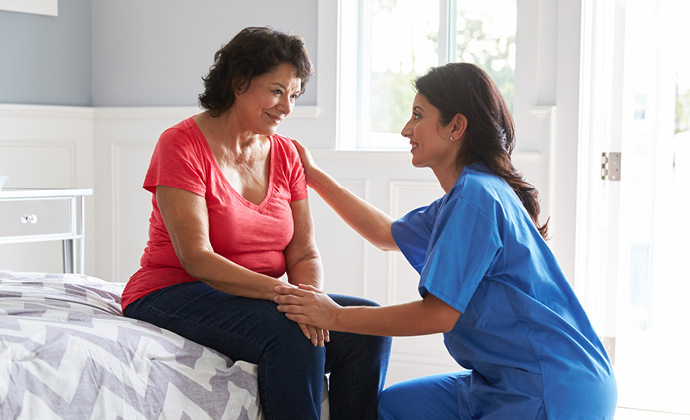A VITAS team member kneels in front of a woman who is sitting at the edge of a bed