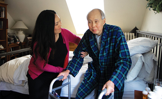 A woman helps her father out of bed with assistance from a walker