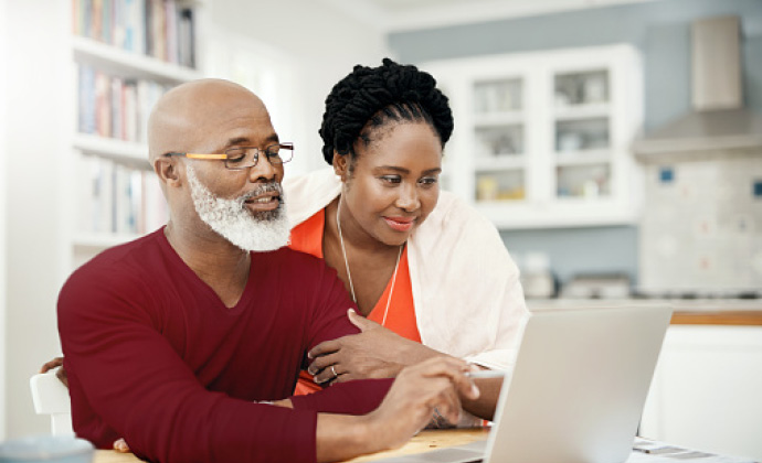 A man uses a laptop at a kitchen table while his wife looks on at the screen